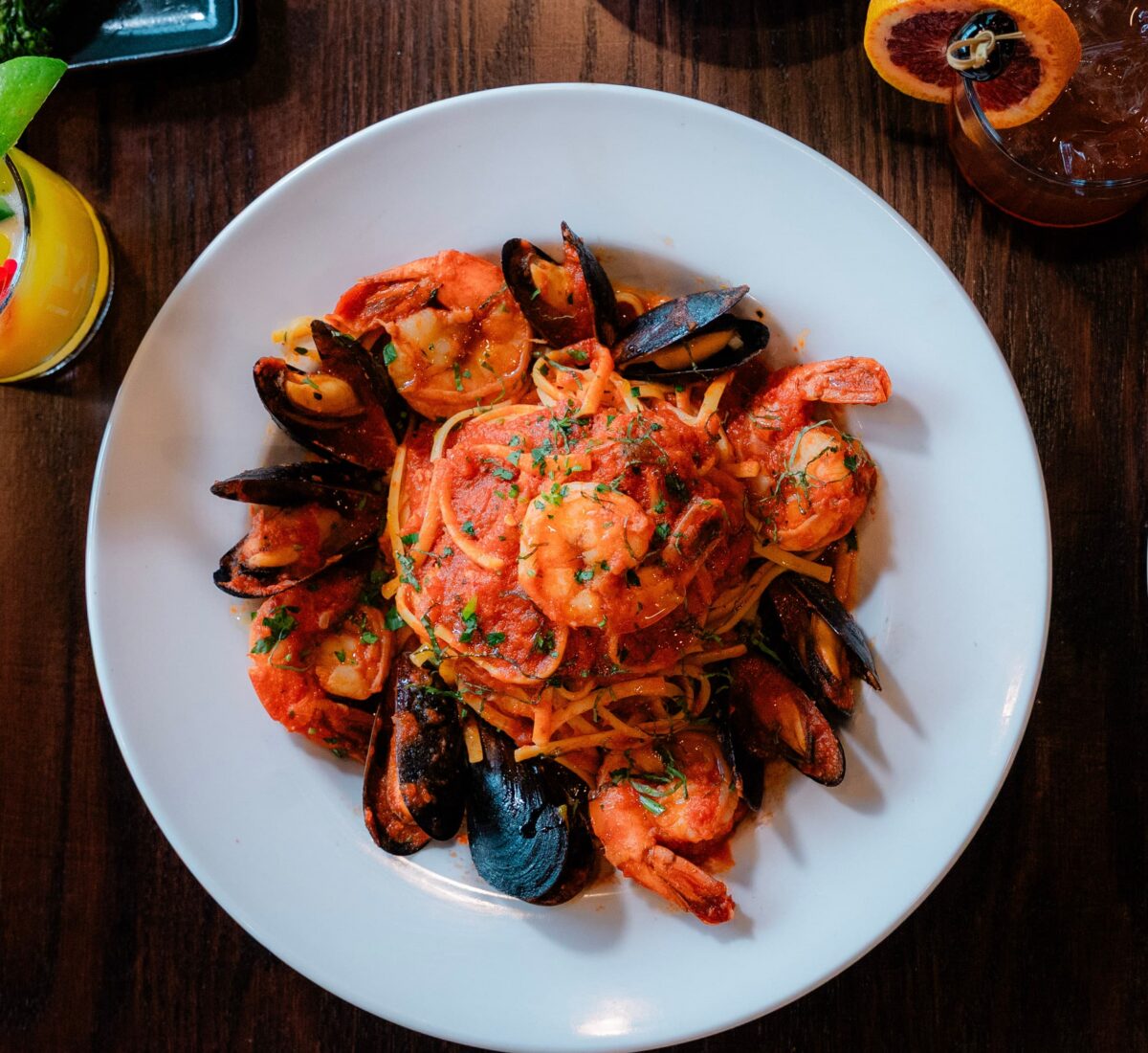 Seafood pasta with shrimp, mussels, and sauce on a white plate, with drinks and side dishes on a wooden table.