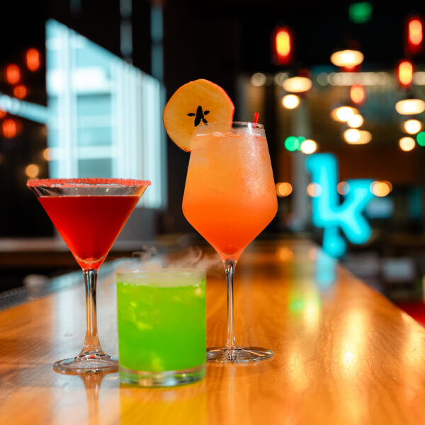 Three colorful cocktails sit on a bar: a red martini, a bright green drink, and an orange drink garnished with an apple slice.