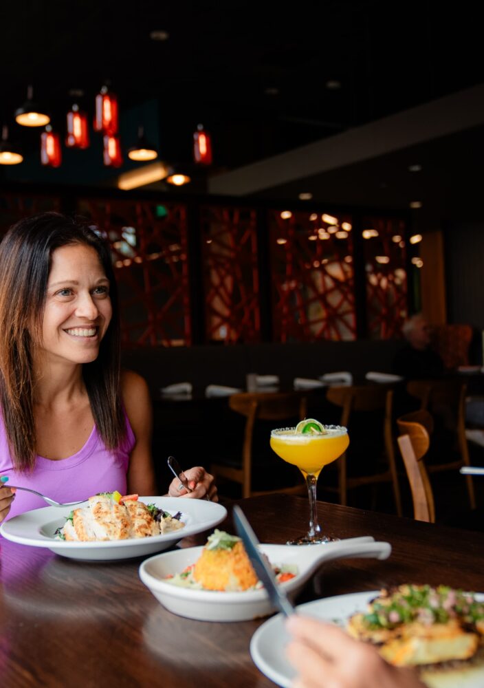 Two women dine together in a restaurant, smiling and enjoying food and drinks, with a modern, warmly lit interior in the background.