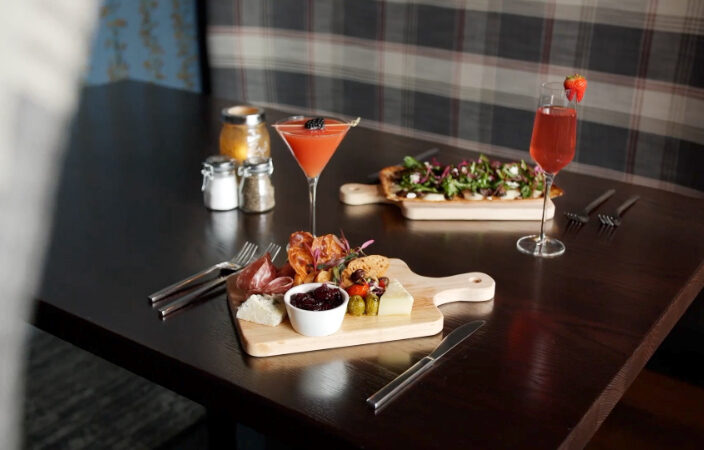 Charcuterie board with meats, cheese, and jam, two cocktails, and a salad pizza on a dark table in a cozy restaurant.