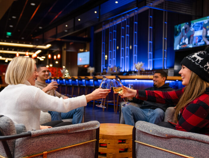 Four people sit indoors, smiling and toasting with wine glasses; a TV is on in the background, and the setting has modern, blue accent lighting.