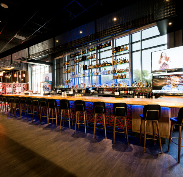 Modern bar with high stools, illuminated counter, liquor bottles on shelves, TV screens, and large windows letting in natural light.