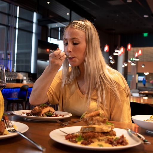 A woman with long blonde hair eats food at a restaurant table, surrounded by plates of meals under warm lighting.