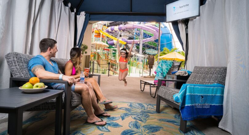 A child jumps joyfully outside a cabana while two adults relax inside, near a water park with colorful slides in the background.