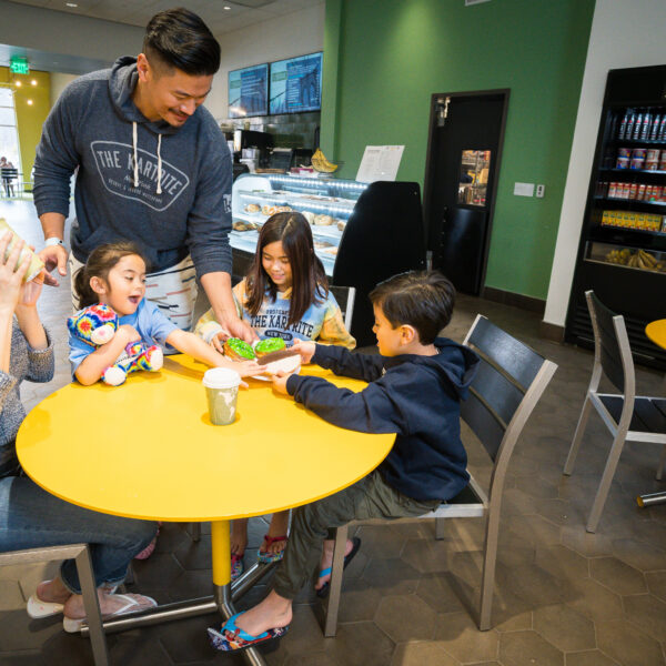 A family of five enjoys drinks and snacks together at a yellow table in a bright, modern café.