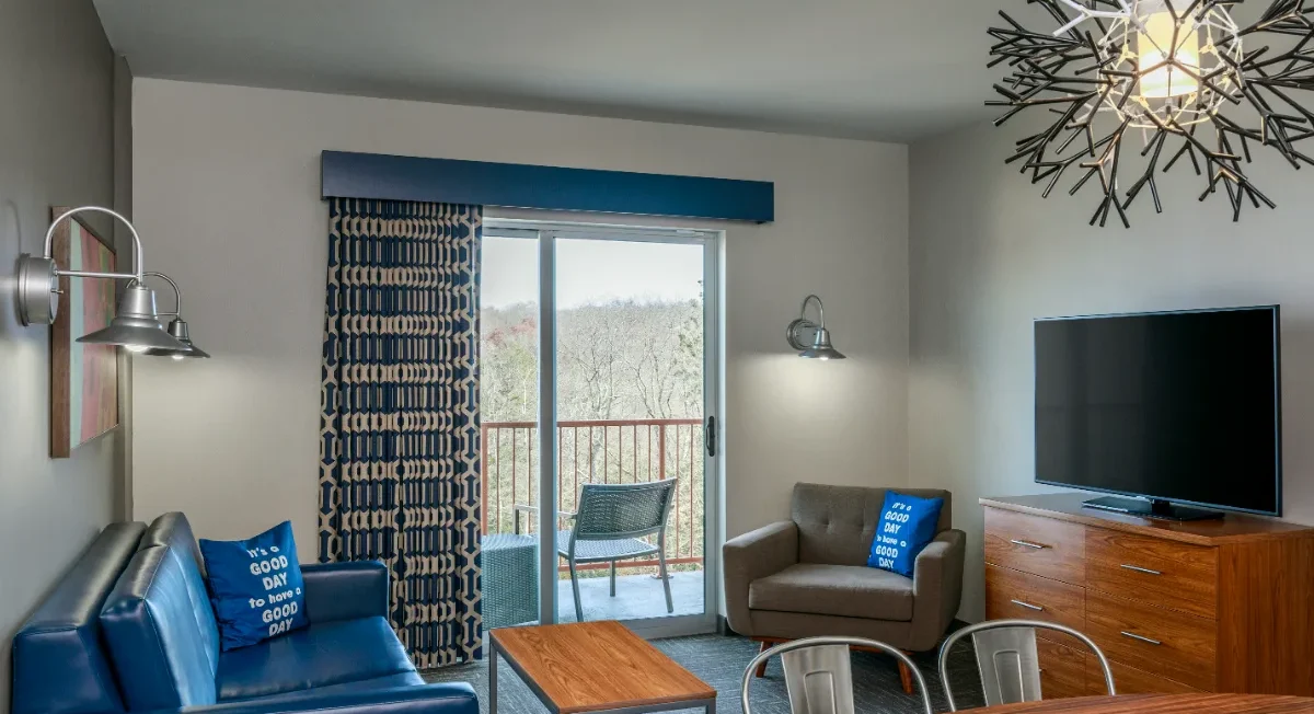Modern living room with blue sofa, armchair, TV, coffee table, balcony with chairs, large window, and decorative lighting fixture.