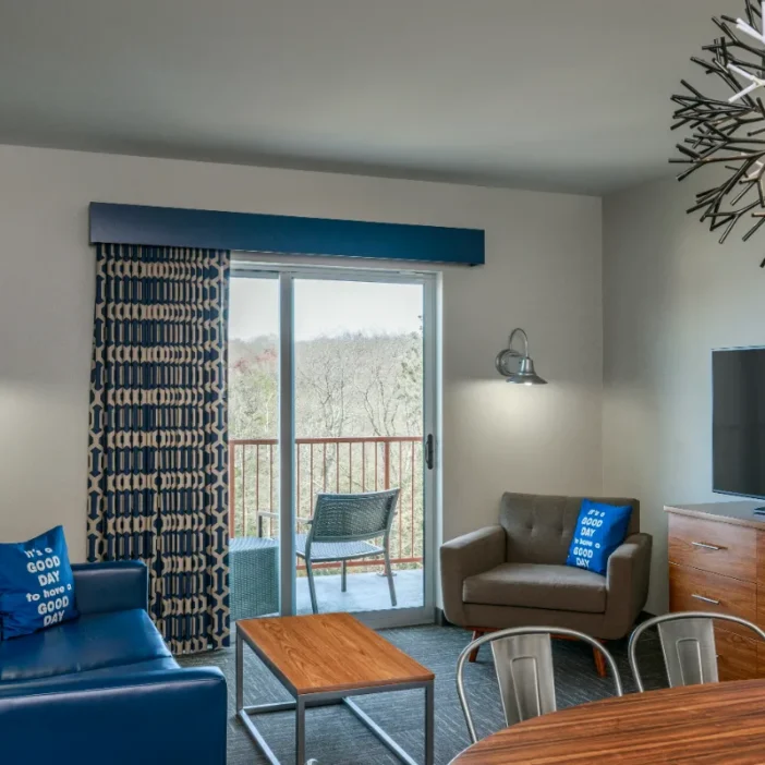 Modern living room with blue sofa, armchair, TV, coffee table, balcony with chairs, large window, and decorative lighting fixture.