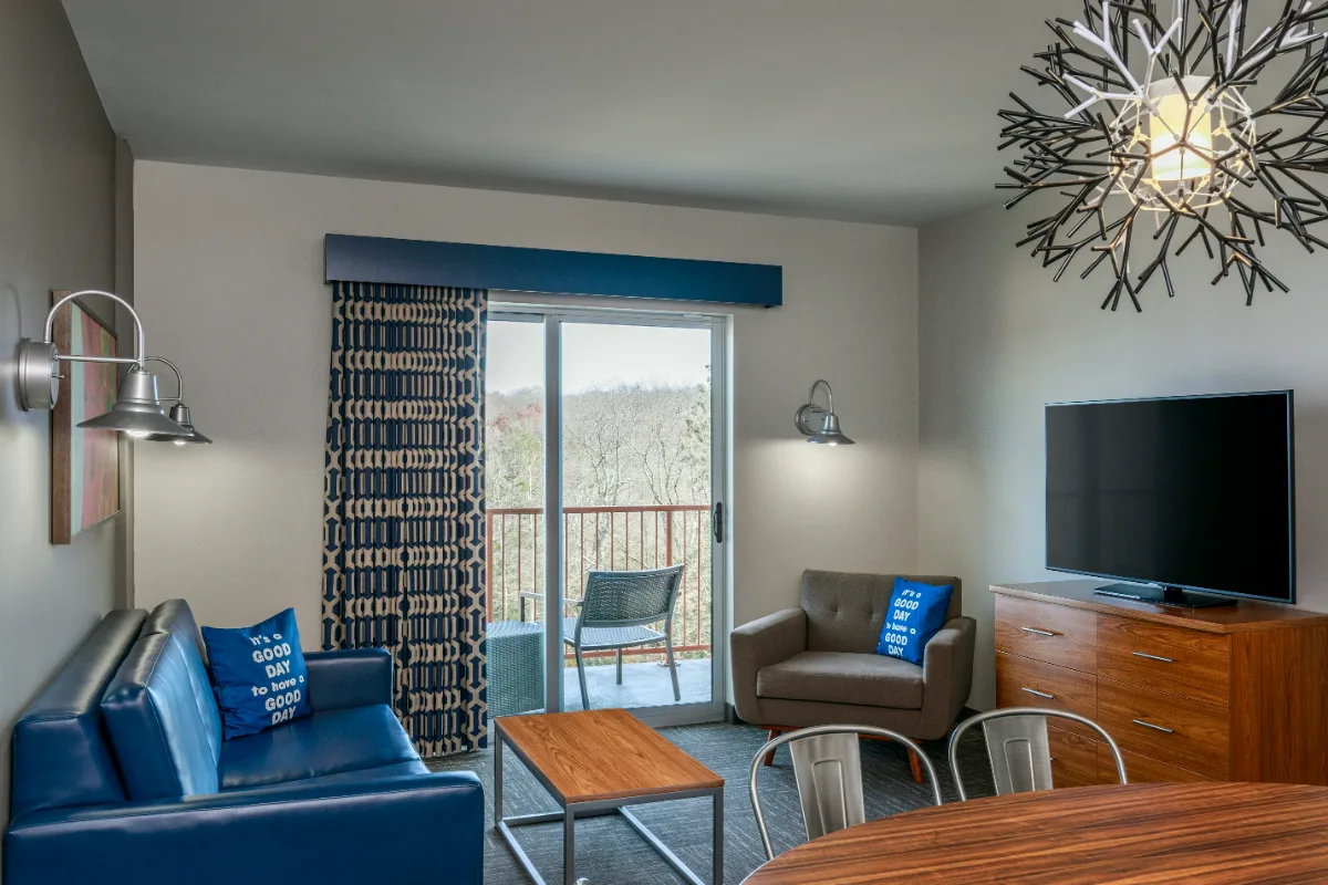 Modern living room with blue sofa, armchair, TV, coffee table, balcony with chairs, large window, and decorative lighting fixture.