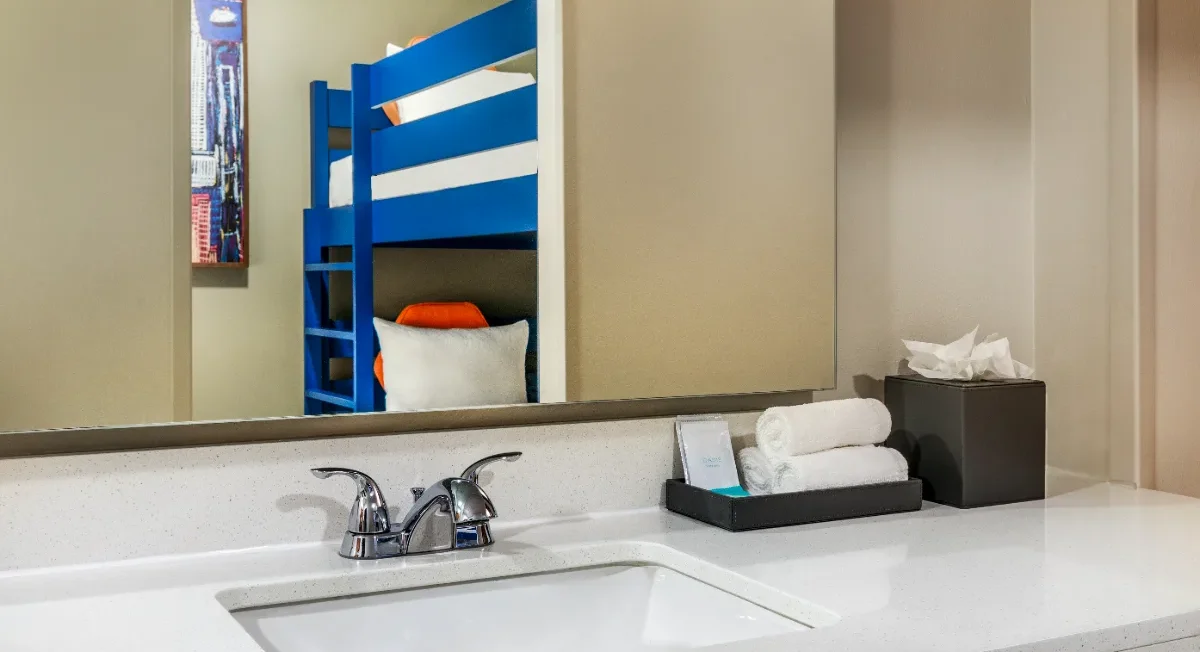A bathroom sink with towels and tissues on the counter, reflected in the mirror is a room with a blue bunk bed and a pillow.