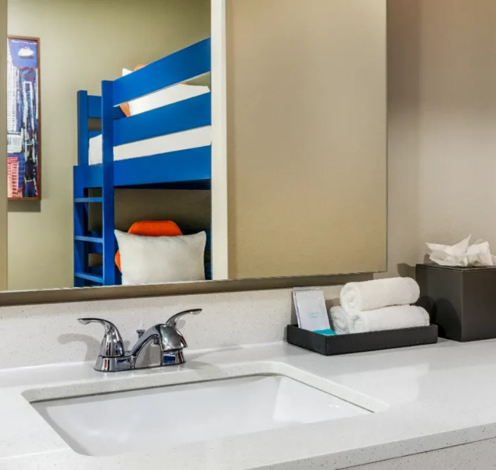 A bathroom sink with towels and tissues on the counter, reflected in the mirror is a room with a blue bunk bed and a pillow.