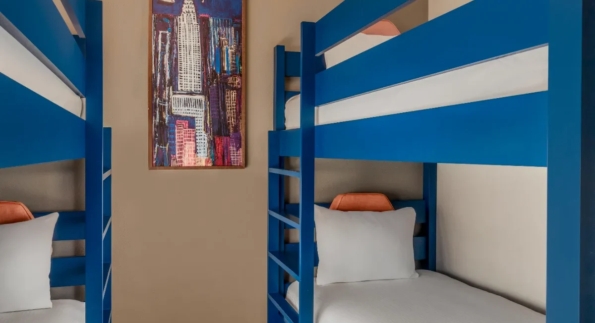 Room with two blue bunk beds, white bedding, and a framed cityscape painting on the beige wall between the beds.