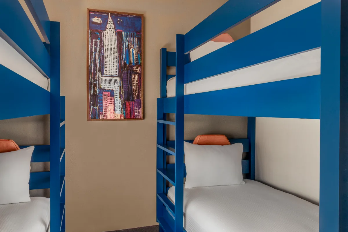 Room with two blue bunk beds, white bedding, and a framed cityscape painting on the beige wall between the beds.