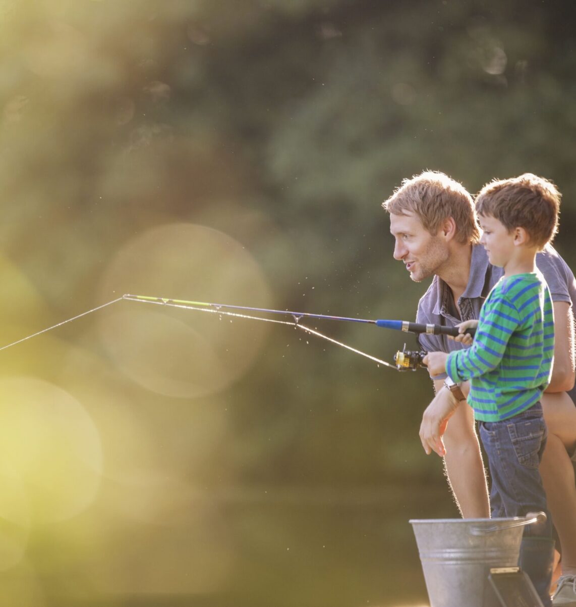 An adult and a child are fishing together outdoors, kneeling by a pond with a fishing pole and a metal bucket nearby.