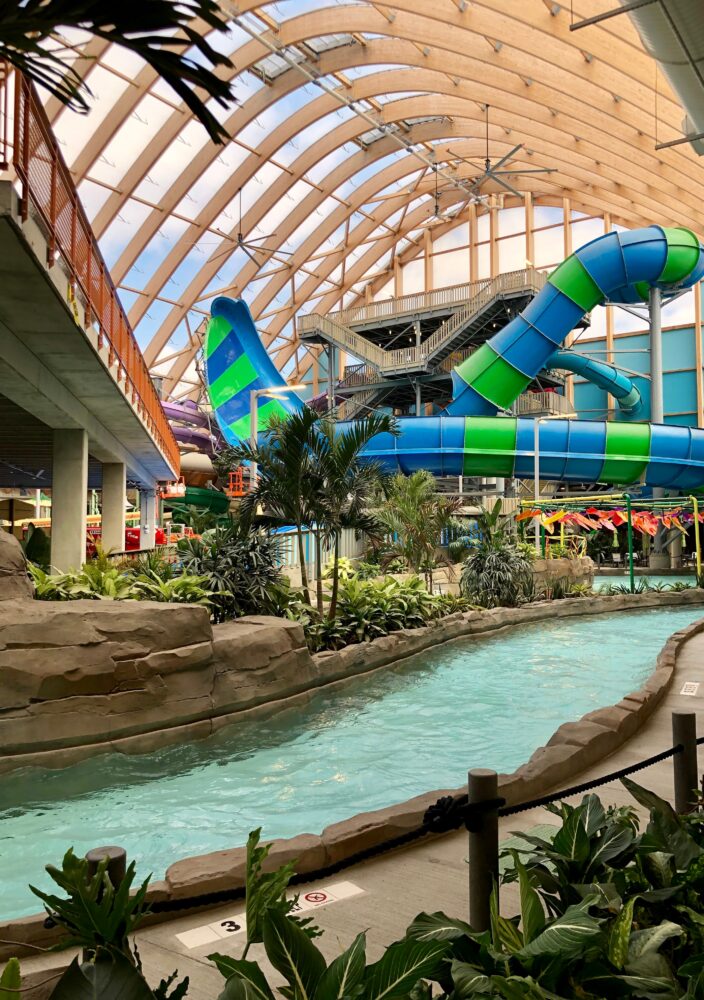 Indoor water park with a lazy river, tropical plants, and large blue and green waterslides under a curved wooden roof.