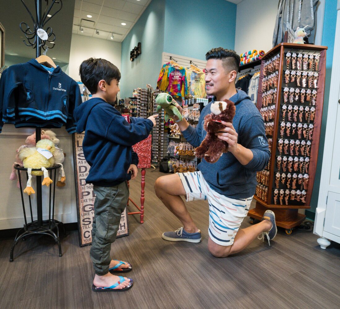 A man kneels, holding a sloth plush toy, as a boy in flip-flops holds out a turtle toy in a gift shop filled with souvenirs and stuffed animals.