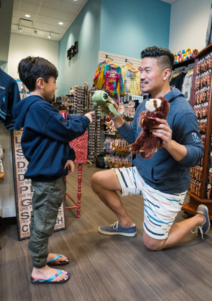 A man kneels, holding a sloth plush toy, as a boy in flip-flops holds out a turtle toy in a gift shop filled with souvenirs and stuffed animals.