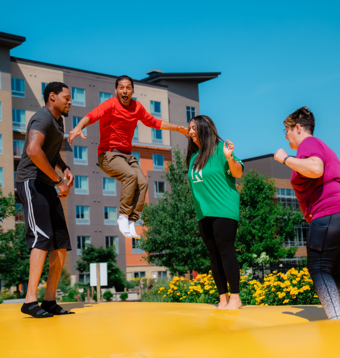 Four adults are laughing and jumping on a large yellow inflatable outside, with buildings and greenery in the background under a clear blue sky.