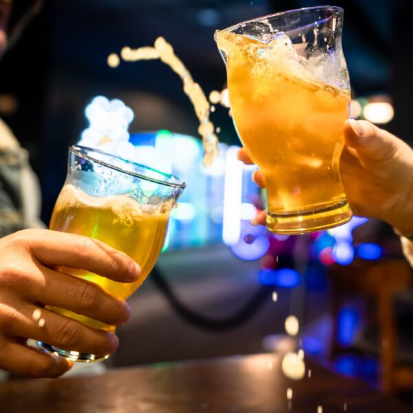 Two people clink glasses of beer, causing some beer to splash out, while sitting at a bar with colorful lights in the background.