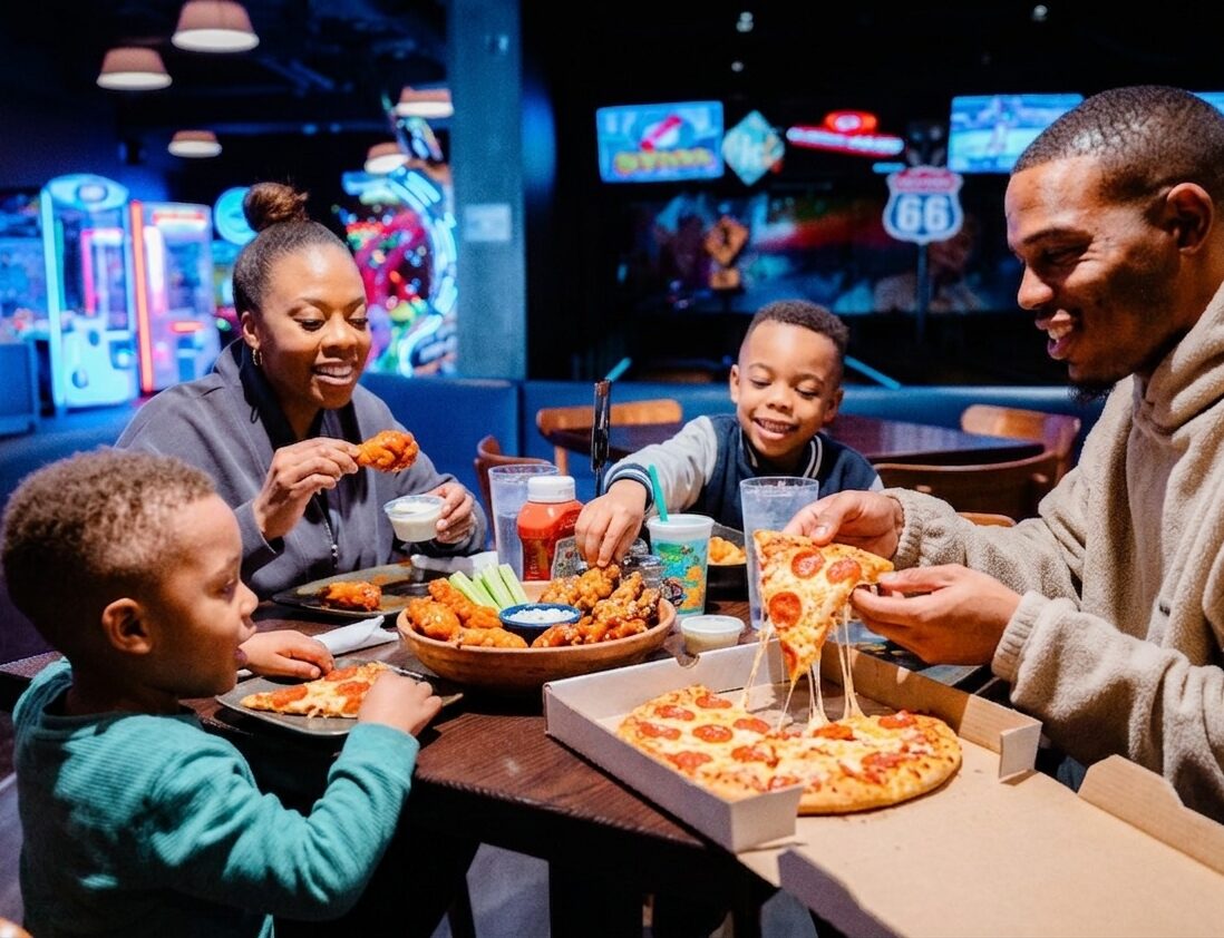 A family of four enjoys pizza, wings, and drinks at a restaurant with arcade games and neon lights in the background.