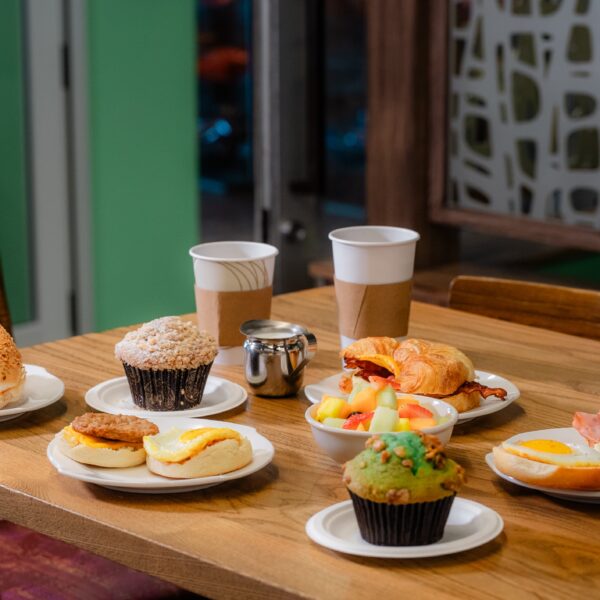 A table with breakfast foods: bagel, muffins, egg sandwiches, fruit, croissant, ham, and two paper coffee cups in a cafe setting.