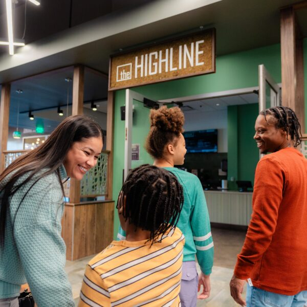Four people smile and walk into a brightly lit café with a sign reading the HIGHLINE above the entrance.