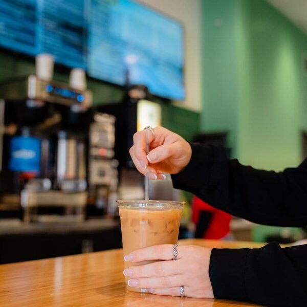 A person stirs iced coffee in a plastic cup on a wooden table inside a café with a green interior and digital menu boards.