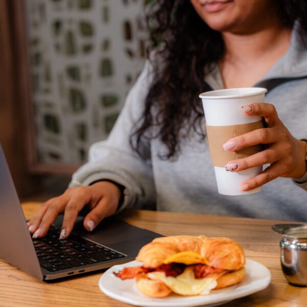 Woman using a laptop at a table, holding a coffee cup, with a croissant sandwich and a small pitcher beside her.