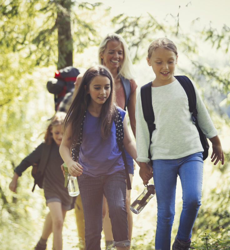A group of children and an adult hike through a sunlit forest, carrying backpacks and water bottles, smiling and enjoying nature.