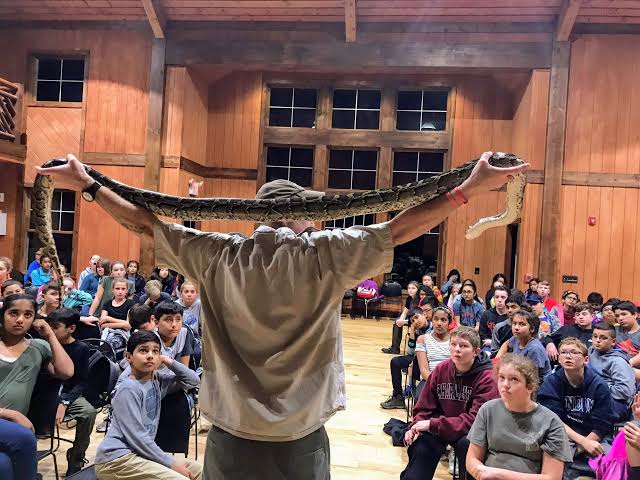 A person holds a large snake over their shoulders in front of a group of children seated in a wooden room, watching attentively.