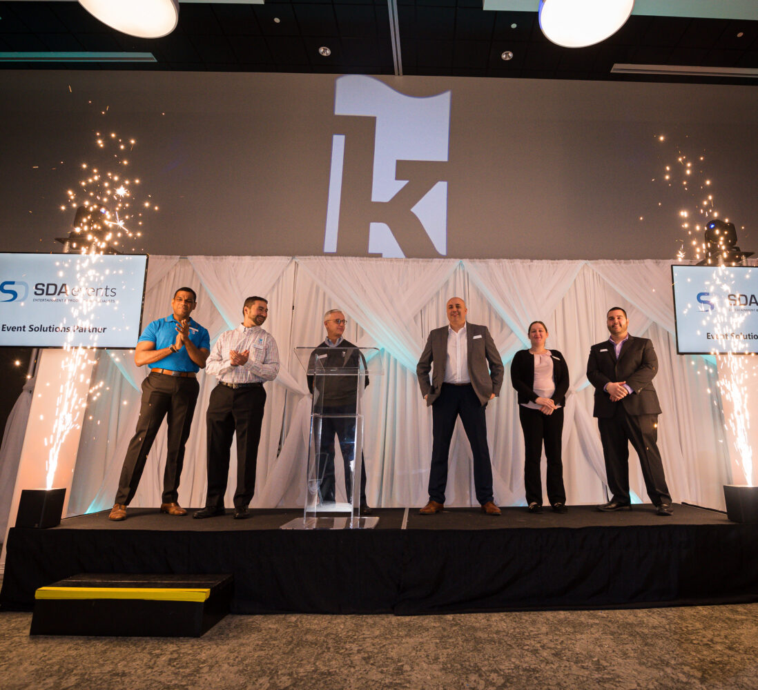 Six people stand on a stage with white drapes and sparklers, two screens display SDA Events, and a large K is illuminated on the wall.