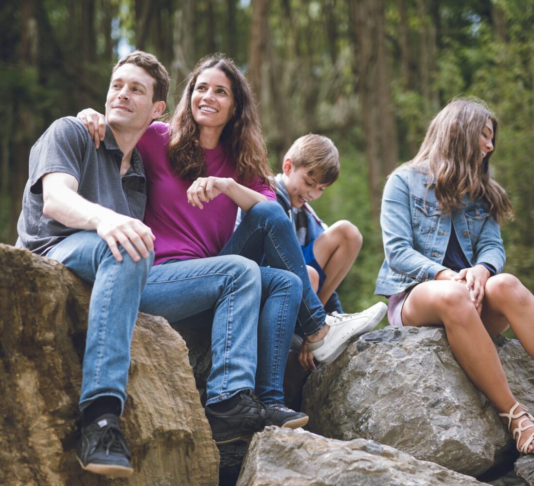 A family of four sits on large rocks outdoors, smiling and relaxing together in a wooded area.