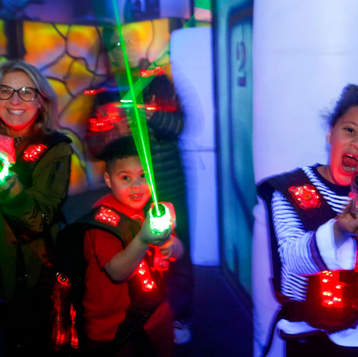 Three people wearing vests play laser tag, smiling and aiming glowing laser guns in a colorful, dimly lit arena.