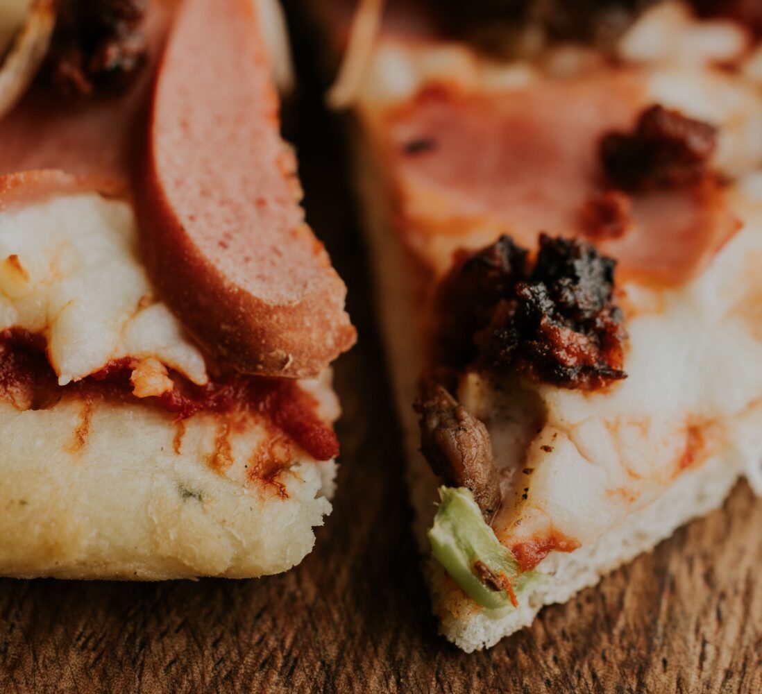 Close-up of two slices of pizza with cheese, ham, sausage, and vegetables on a wooden surface.