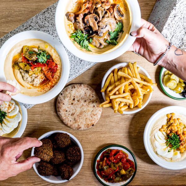 A table with pita bread, hummus, falafel, fries, vegetables, olives, and two people reaching for food. Mediterranean meal is being shared.