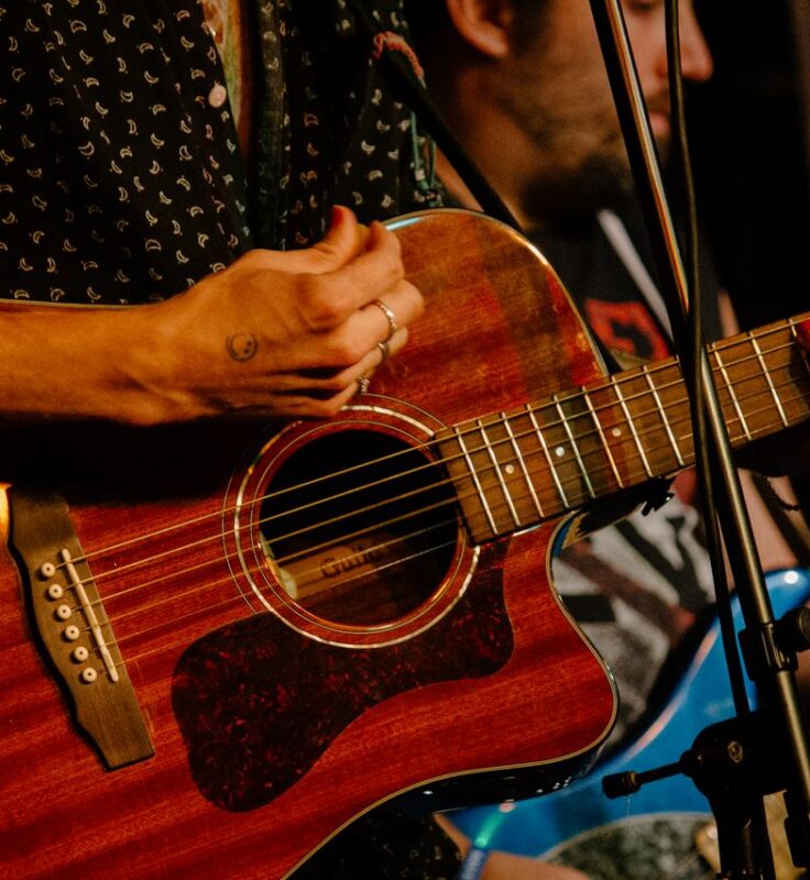 Two musicians playing guitar, one with an acoustic guitar in the foreground and one with an electric guitar in the background, both performing on stage.