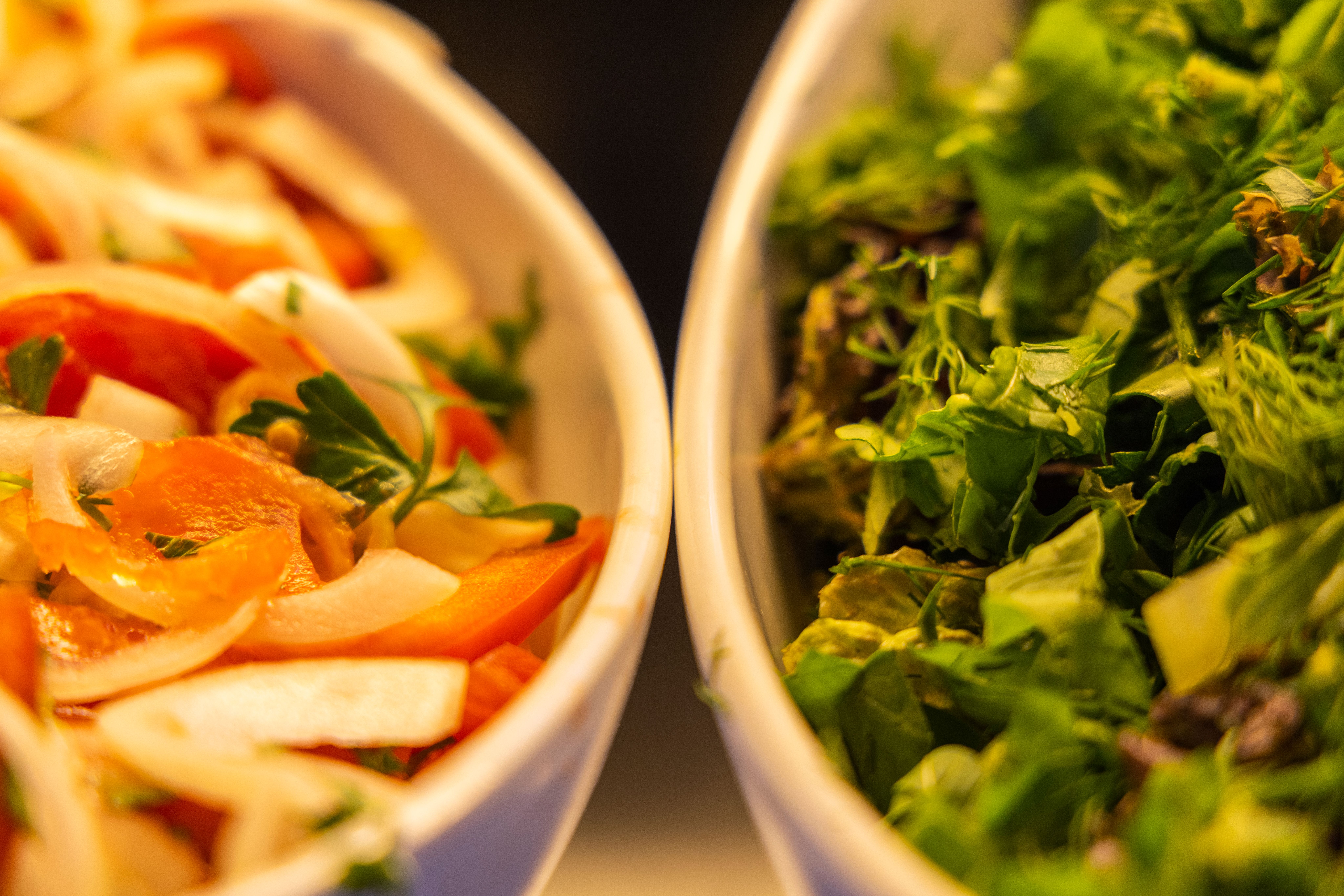 Close-up of two bowls: one with sliced tomatoes and onions, the other with chopped leafy greens, both arranged in white dishes.