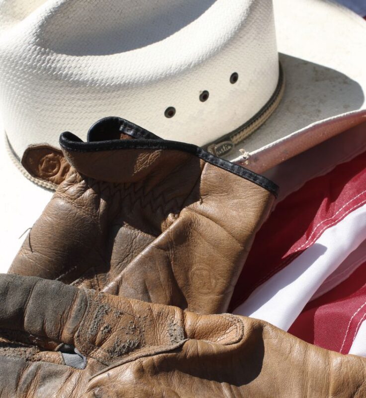 A white cowboy hat and worn brown gloves rest on an American flag, symbolizing western culture and patriotism.