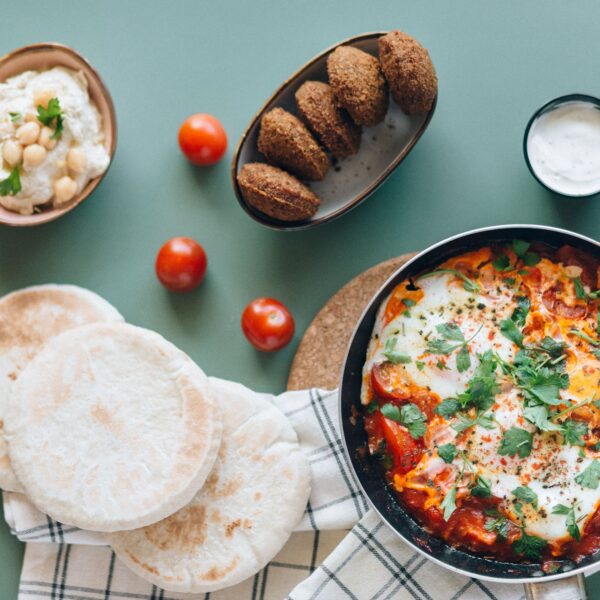 A table with pita bread, shakshuka in a pan, falafel, hummus, cherry tomatoes, and a small bowl of white sauce.