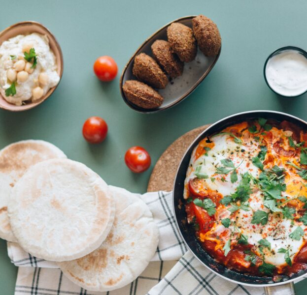 A table with pita bread, shakshuka in a pan, falafel, hummus, cherry tomatoes, and a small bowl of white sauce.