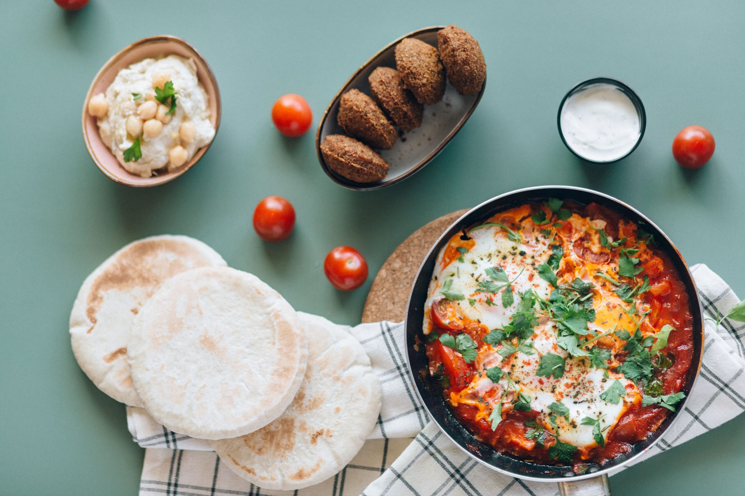 A table with pita bread, shakshuka in a pan, falafel, hummus, cherry tomatoes, and a small bowl of white sauce.