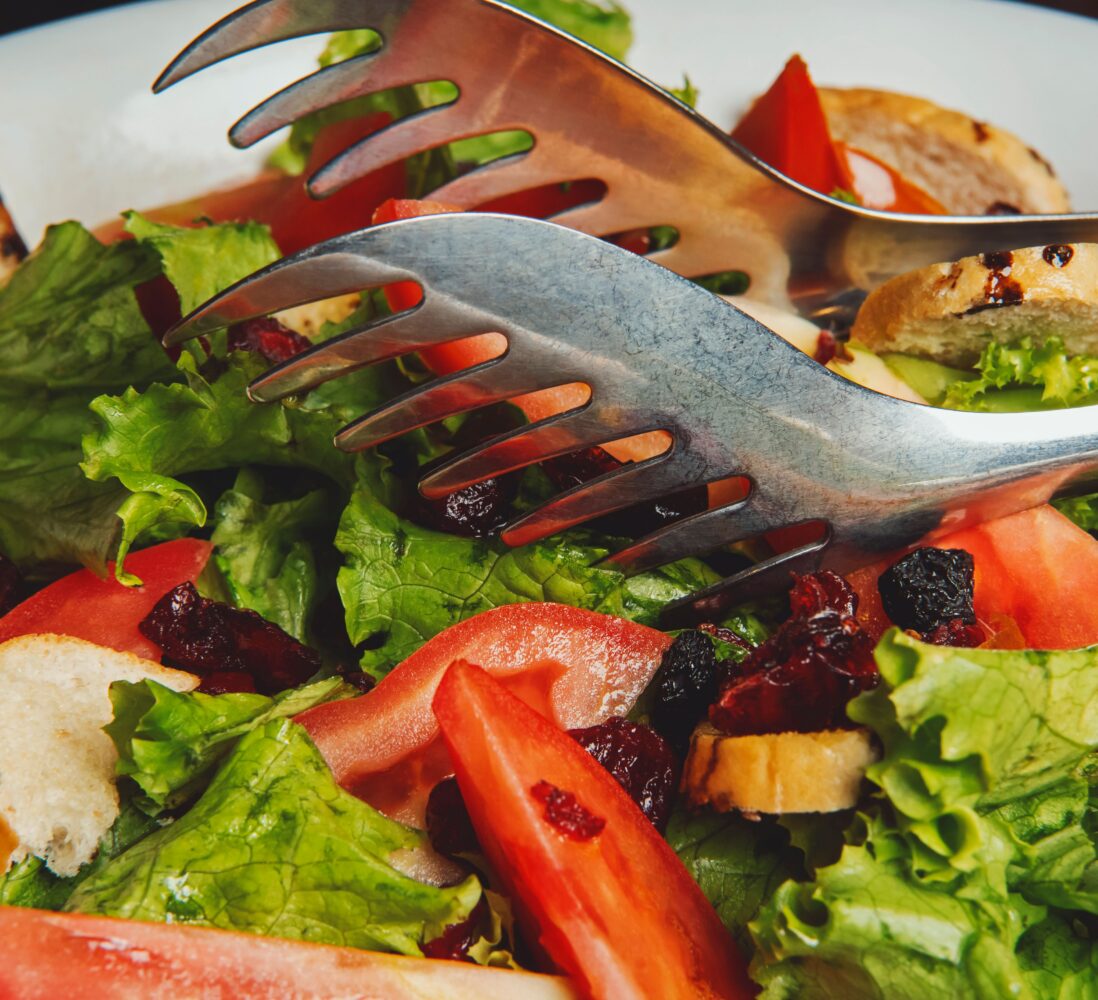 A bowl of fresh salad with lettuce, tomato wedges, dried cranberries, croutons, and metal salad tongs resting on top.