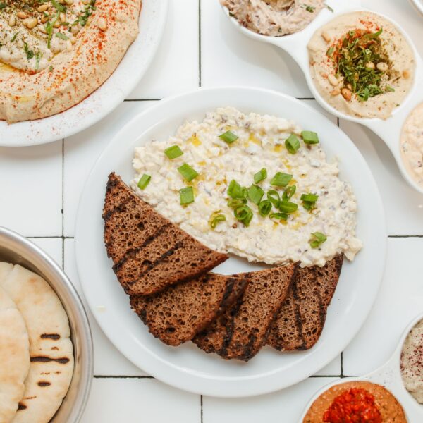 Plate of dark bread slices with creamy dip topped with green onions, surrounded by hummus, pita bread, and various dips on a white tiled table.