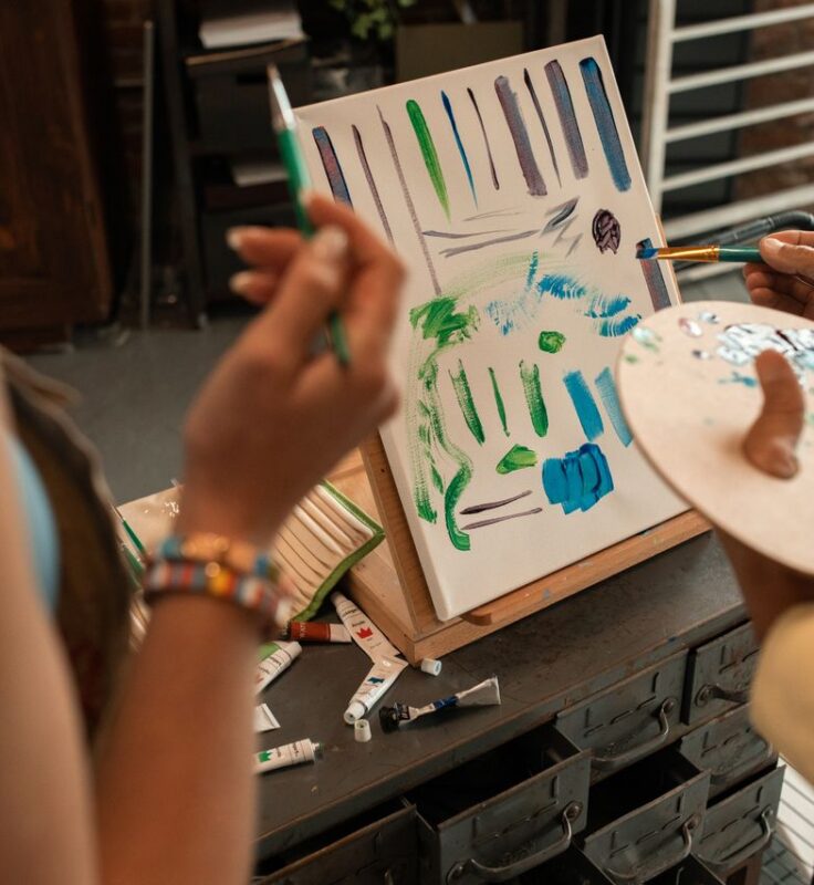 Two people paint an abstract image with green and blue brushstrokes on a canvas, using brushes and a palette in a bright indoor studio.
