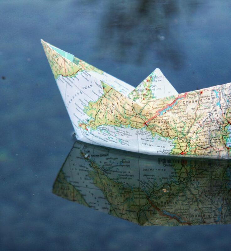 A paper boat made from a world map floats on calm water, reflecting its image on the surface.