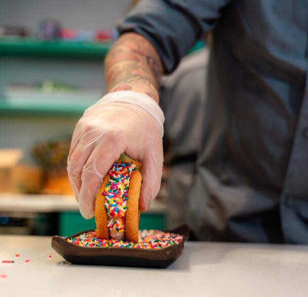 A person in gloves dips an ice cream taco with sprinkles into a plate of colorful sprinkles on a counter.