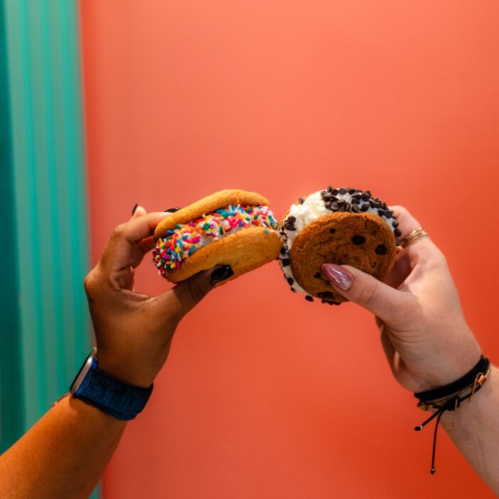 Two hands hold up different ice cream sandwiches with colorful sprinkles and chocolate chips against a coral and teal background.