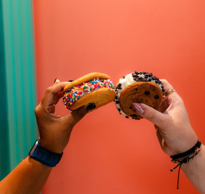 Two hands hold up different ice cream sandwiches with colorful sprinkles and chocolate chips against a coral and teal background.