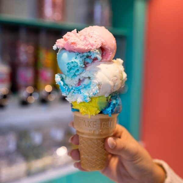A hand holds a colorful ice cream cone with scoops of blue, pink, white, and yellow in a candy shop.