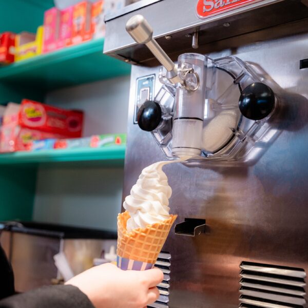 Person filling a waffle cone with vanilla soft serve ice cream from a machine in a colorful shop.