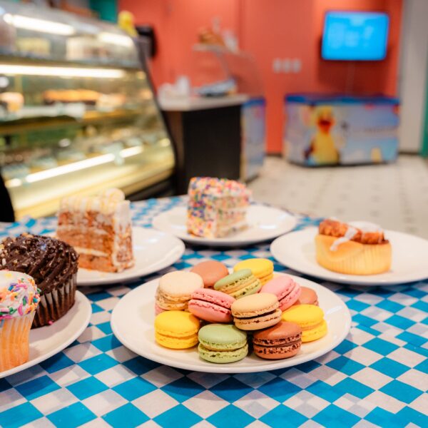 Plates of colorful macarons, cupcakes, and slices of cake are displayed on a blue checkered table in a bakery.